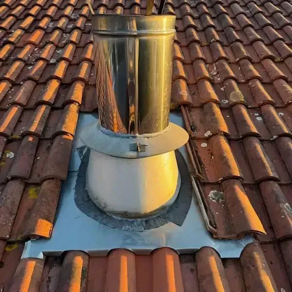 Conduit de cheminée métallique sur un toit de tuiles en terre cuite rouge, avec solin à la base.