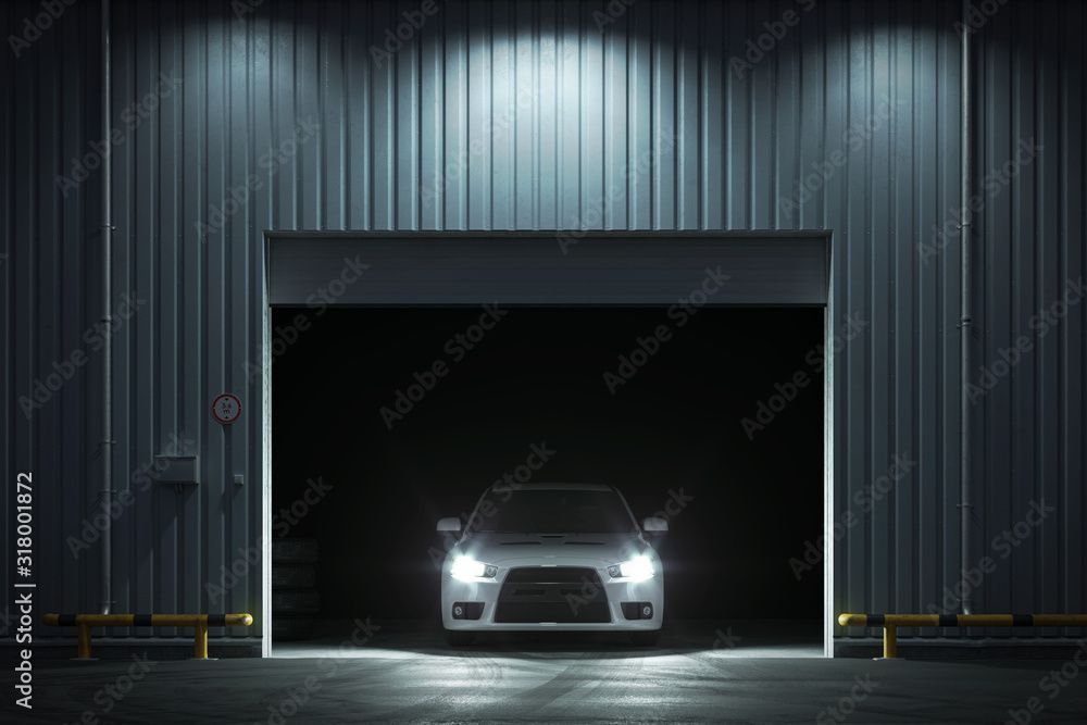 Voiture blanche, phares allumés, dans un garage sombre à porte ouverte, éclairé par des plafonniers.