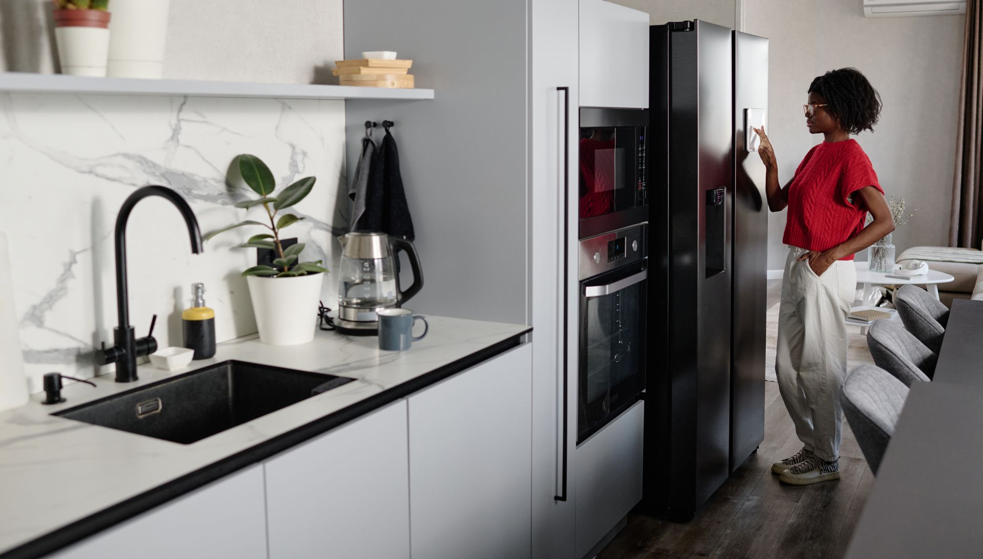 Une personne en haut rouge ouvre un réfrigérateur dans une cuisine moderne avec un évier noir et des placards blancs.