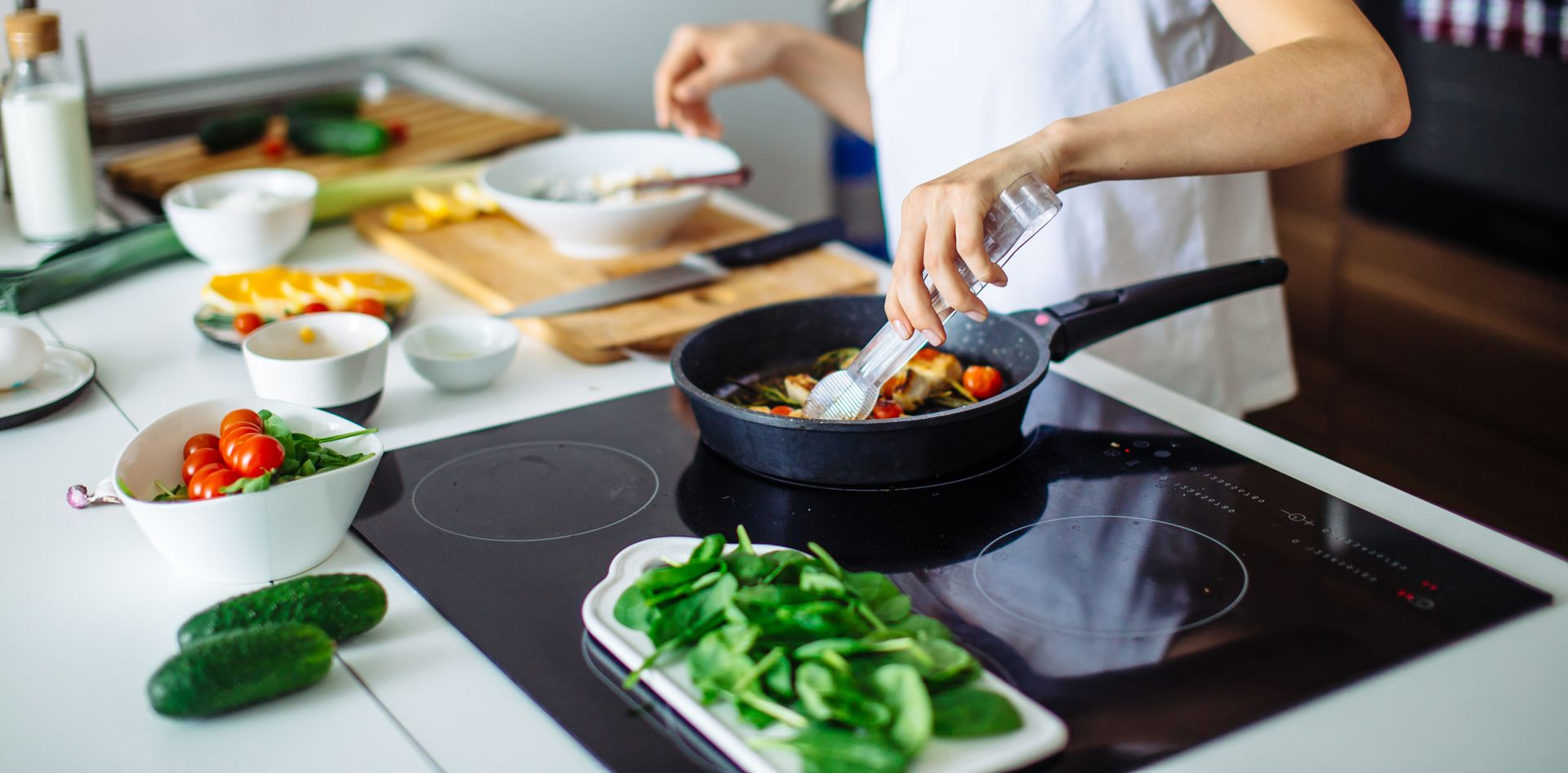 Une personne fait cuire des légumes dans une poêle sur une plaque de cuisson, avec des légumes verts et des tomates fraîches à proximité.