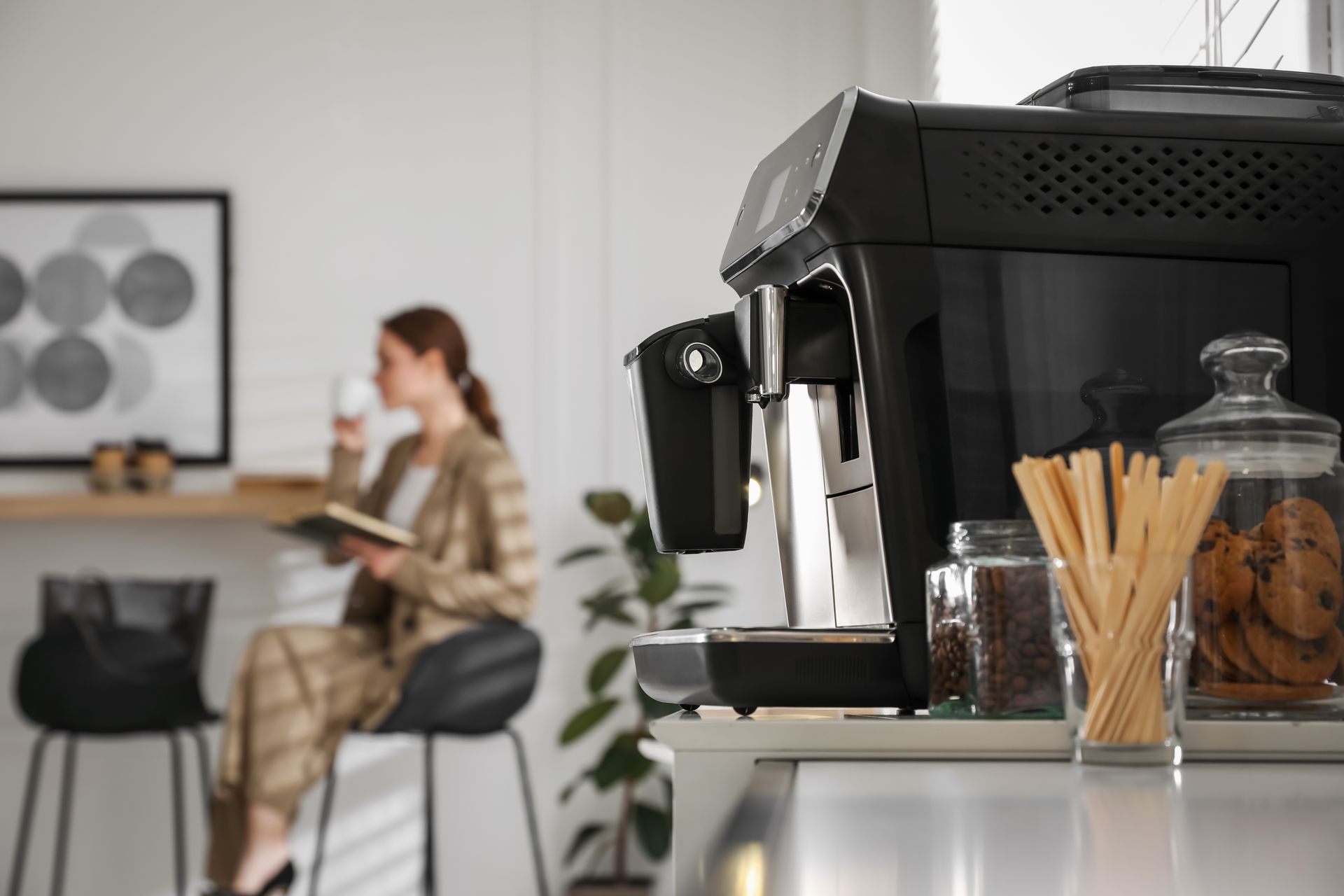 Machine à café au premier plan, une personne assise travaillant dans un bureau lumineux en arrière-plan