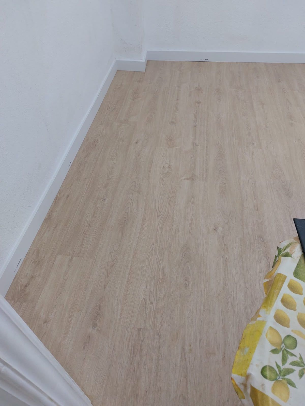 Habitación vacía con suelo de madera clara y paredes blancas; en una esquina se ve parcialmente un bolso floral