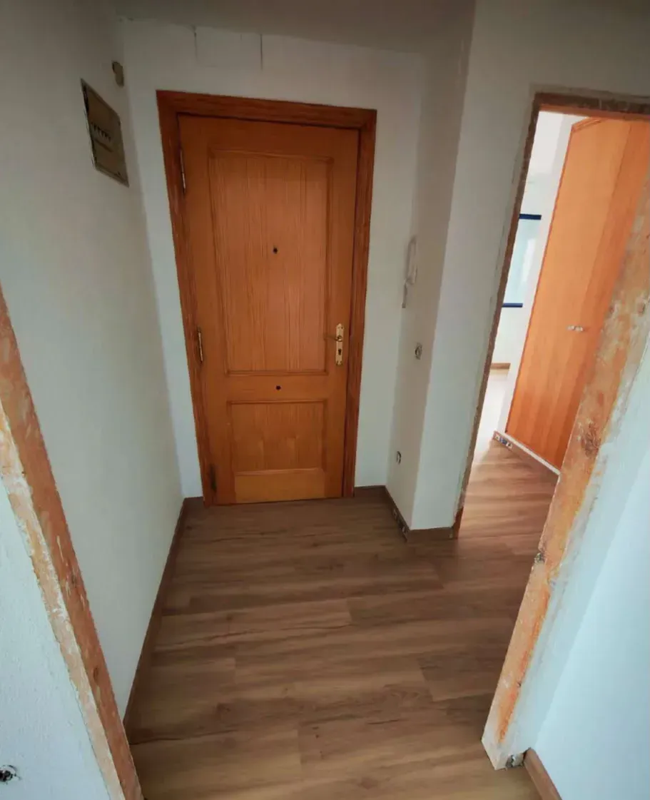 Pasillo interior con una puerta principal de madera y paredes claras, visto desde una puerta abierta.