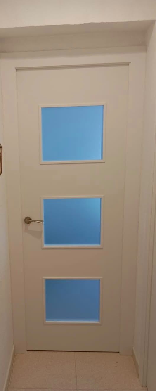 Puerta interior blanca con tres paneles cuadrados de vidrio esmerilado y un tirador metálico.