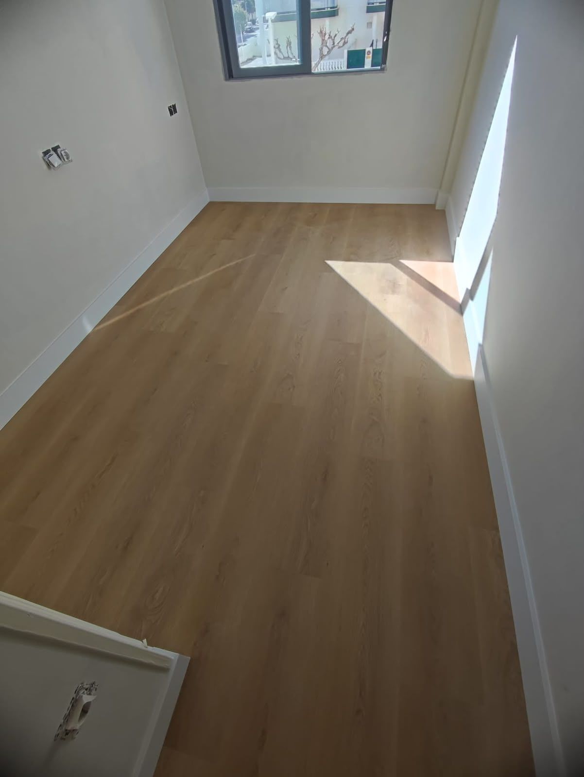 Habitación estrecha y vacía, con suelo de madera clara, paredes blancas y luz del sol junto a la ventana.