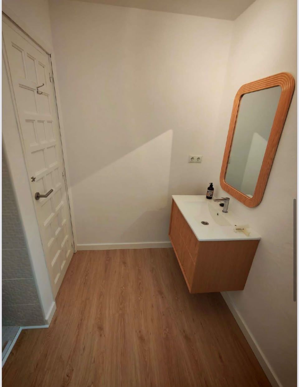 Baño pequeño y blanco con suelo de madera, lavabo con mueble y armario con espejo junto a una puerta.