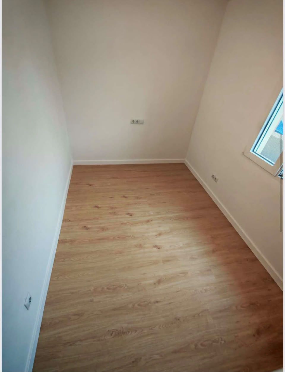 Habitación pequeña vacía con suelo de madera clara, paredes blancas y una ventana a la derecha.