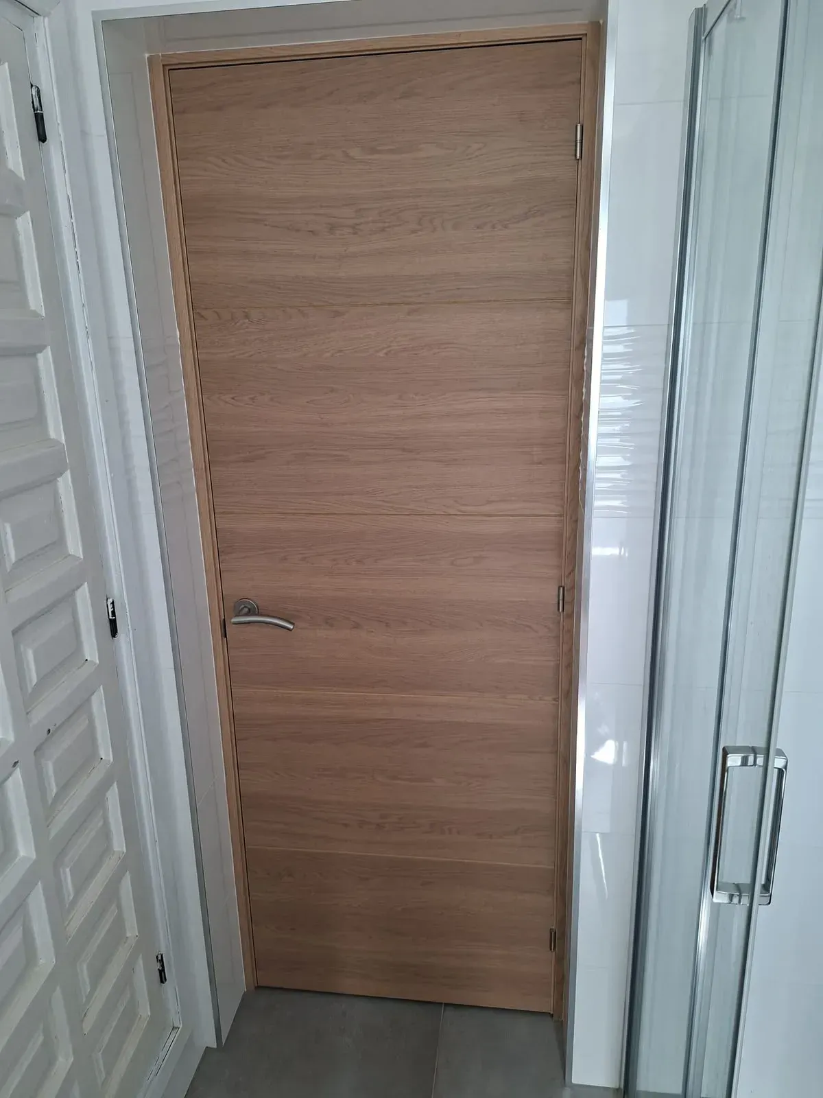 Puerta de madera en un pasillo estrecho y blanco, con una mampara de ducha de cristal a la derecha.