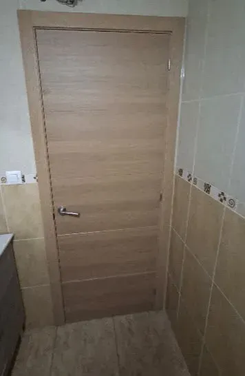 Puerta de baño de madera clara con manilla metálica, enmarcada por paredes de azulejos beige.