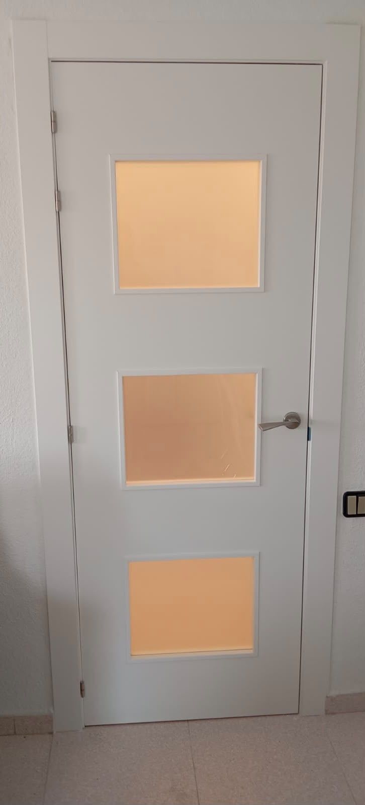 Puerta interior blanca con tres paneles de vidrio esmerilado y un tirador plateado.