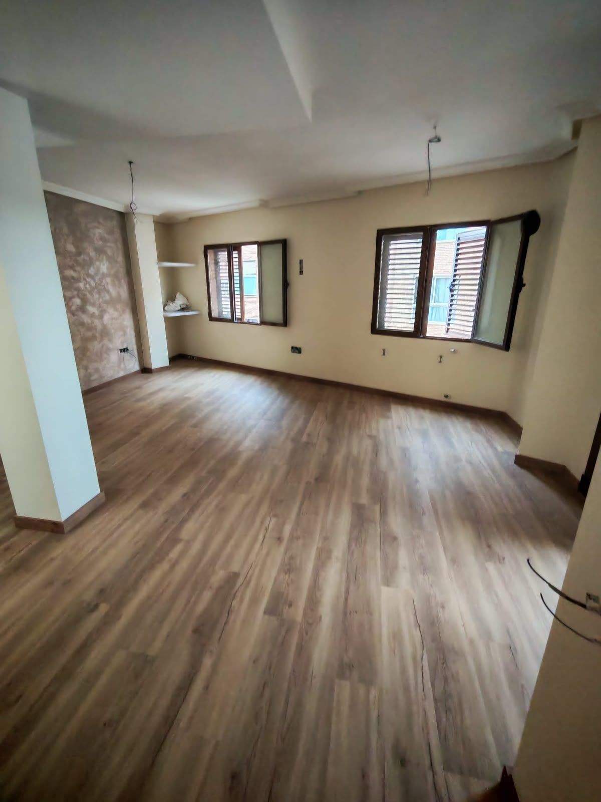 Habitación vacía con suelo de imitación madera, paredes color crema, ventanas y luces de techo a la vista