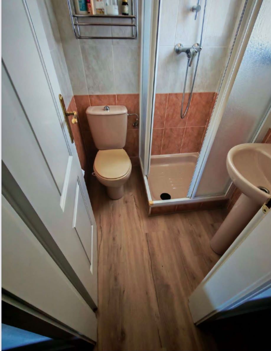 Baño estrecho con inodoro, ducha y lavabo, con suelo de aspecto madera y paredes blancas