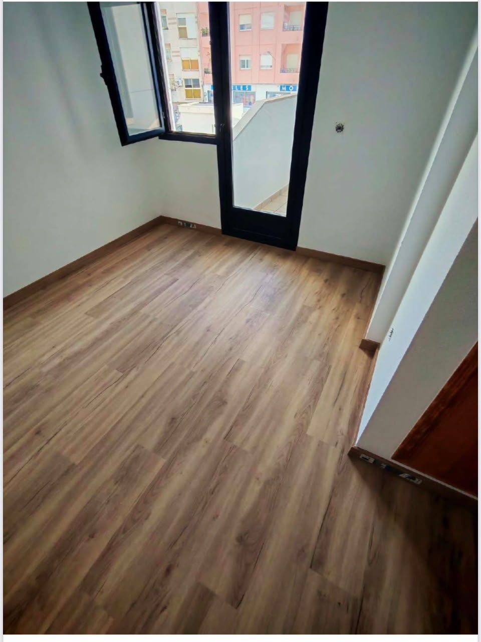 Habitación vacía con suelo de madera y una puerta de cristal con marco negro que da a un balcón.