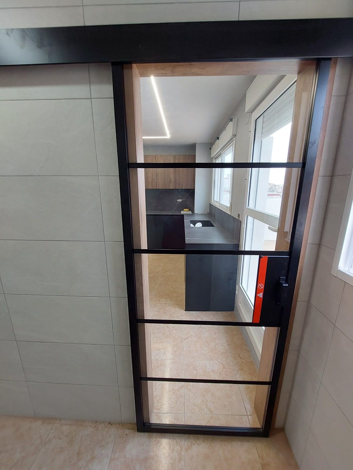 Pasillo interior con una verja de metal negro junto a una pared y una luminosa zona de oficinas con ventanas al fondo
