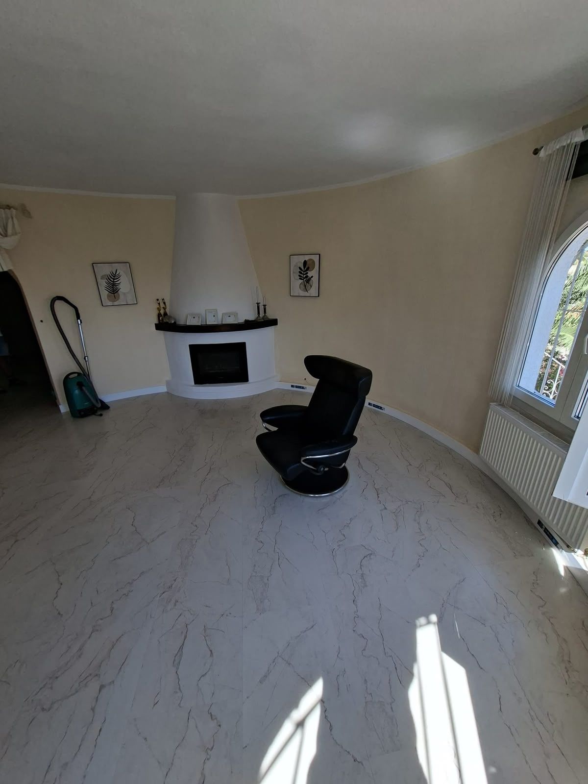 Sala de estar vacía con suelo de baldosas blancas, chimenea, silla negra y un gran ventanal por el que entra la luz del sol