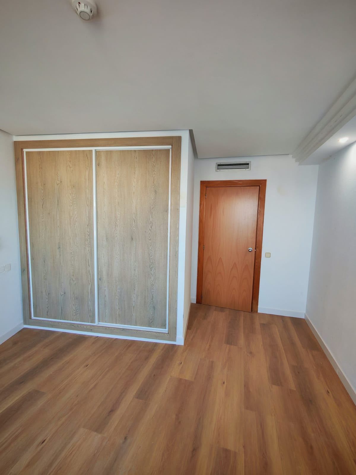 Dormitorio vacío con suelo de madera, puertas correderas en el armario y una puerta de madera en la pared del fondo.