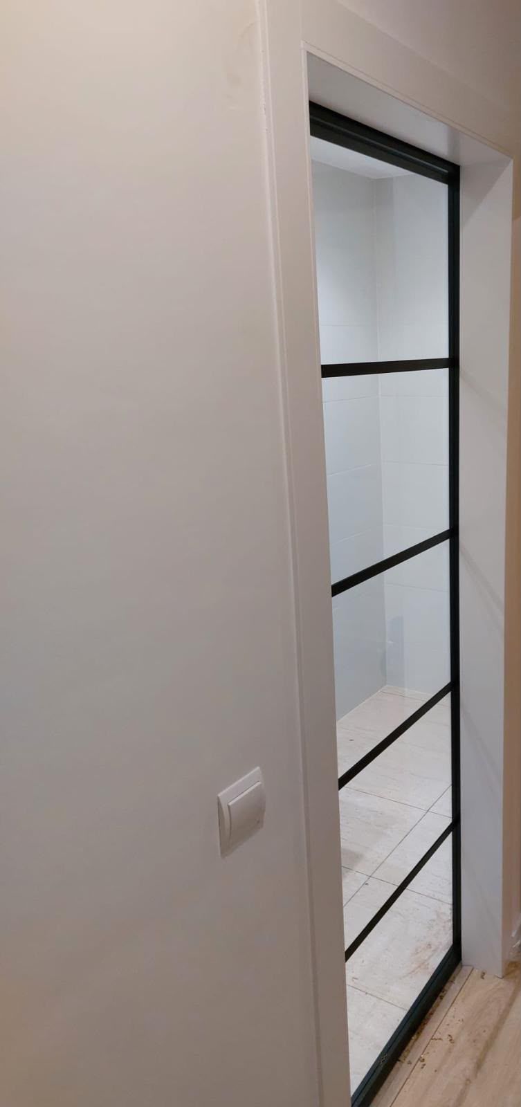 Pasillo estrecho con una puerta de cristal enmarcada en negro y un interruptor blanco en la pared de la izquierda.