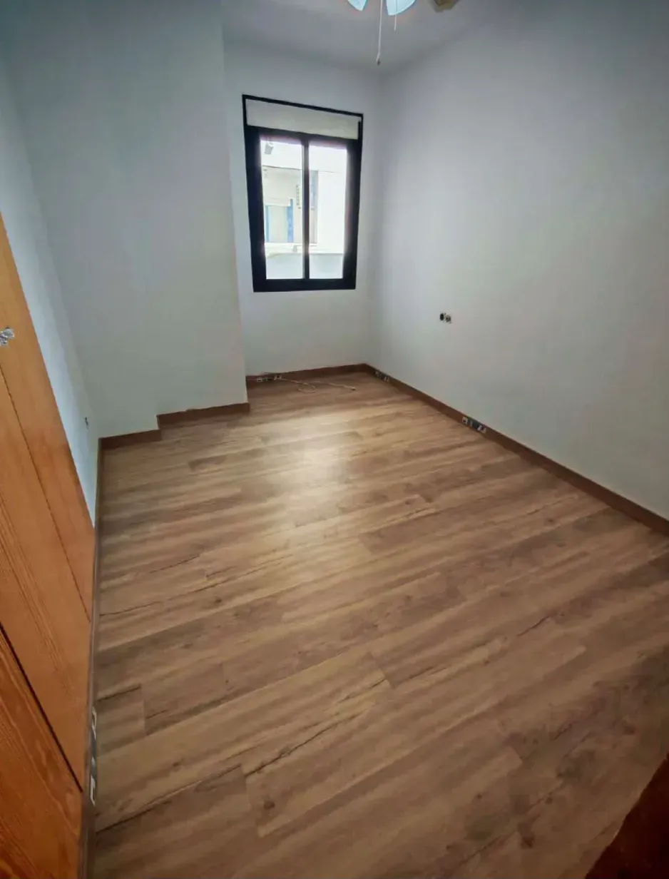 Habitación pequeña y vacía, con suelo de aspecto de madera, paredes blancas y una ventana al fondo.