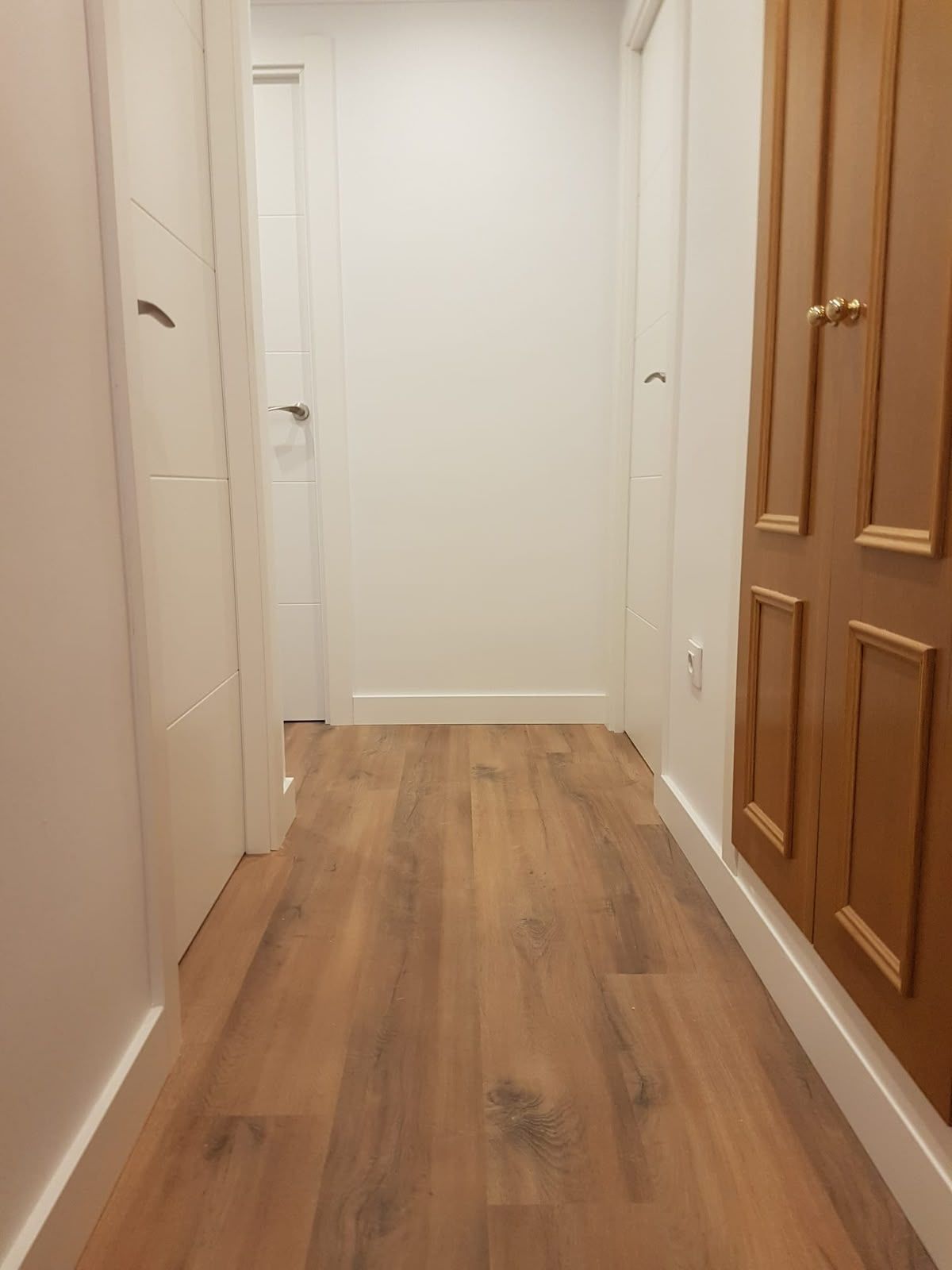 Pasillo estrecho con suelo de aspecto madera, paredes blancas y una puerta de madera a la derecha.