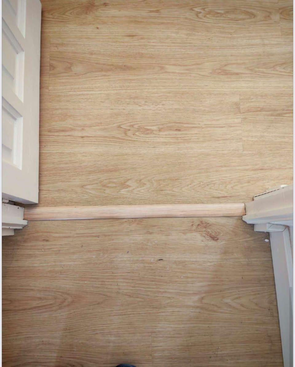 Pasillo estrecho con suelo de madera clara, molduras blancas y un umbral bajo entre dos habitaciones