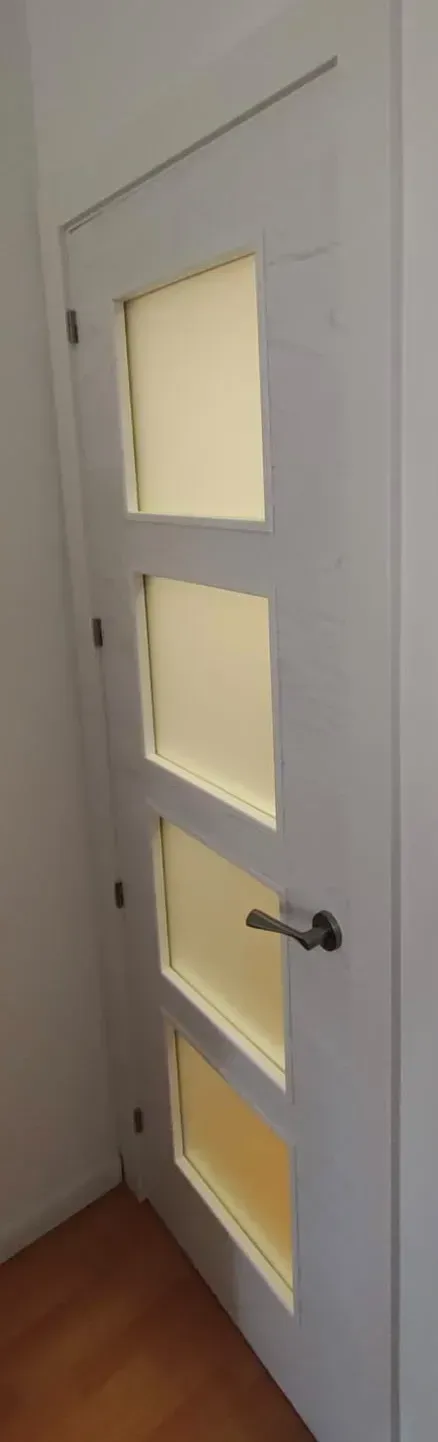 Puerta interior blanca con cuatro paneles de vidrio esmerilado y un picaporte plateado, vista en ángulo.