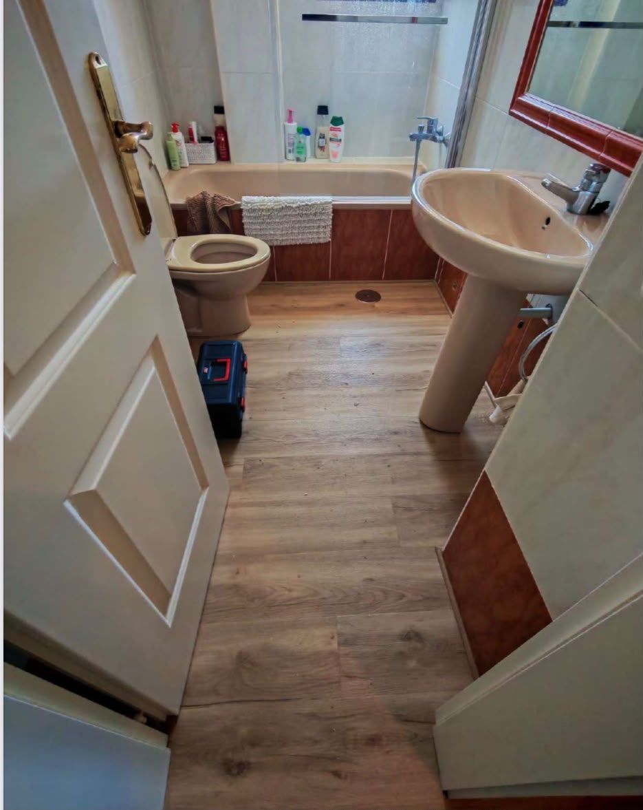Baño pequeño con suelo de imitación madera, lavabo blanco, inodoro, bañera y cortina de ducha.