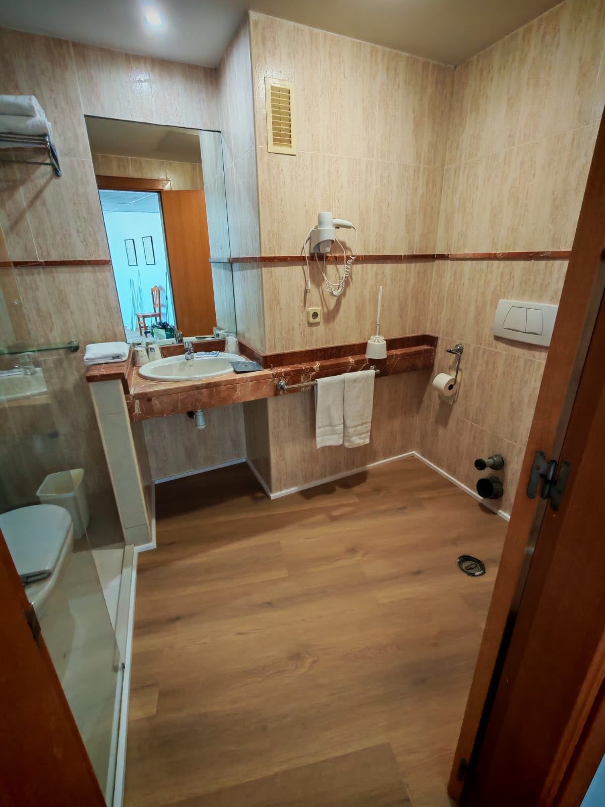 Baño pequeño con tocador de madera, espejo, paredes beige, toallero y ducha a ras de suelo.