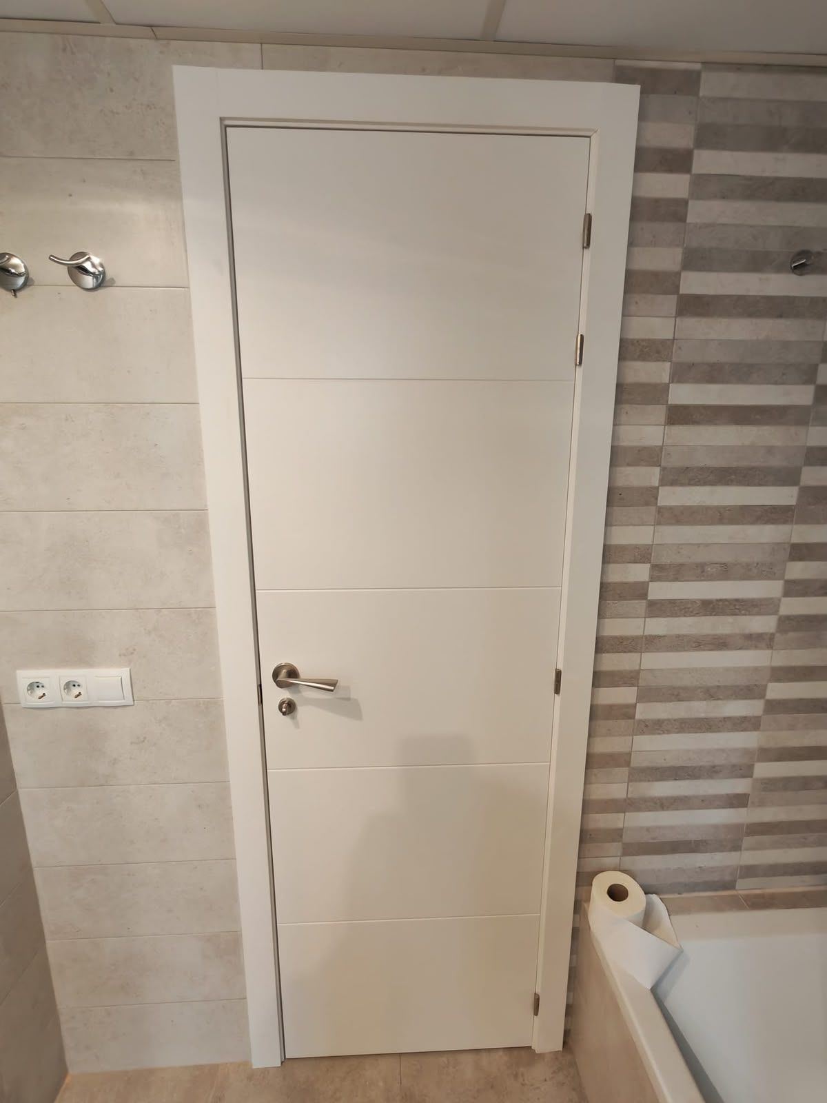 Puerta del baño con paneles blancos, manija plateada, paredes alicatadas y borde de la bañera a la vista.