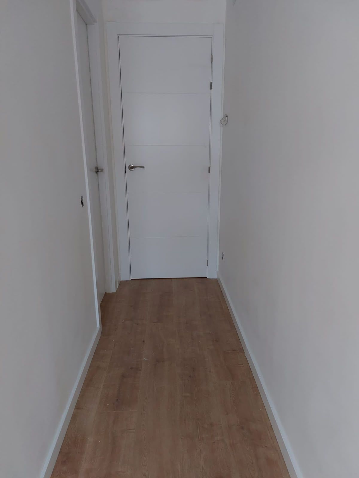 Pasillo estrecho y blanco, con suelo de madera, que conduce a una puerta blanca cerrada