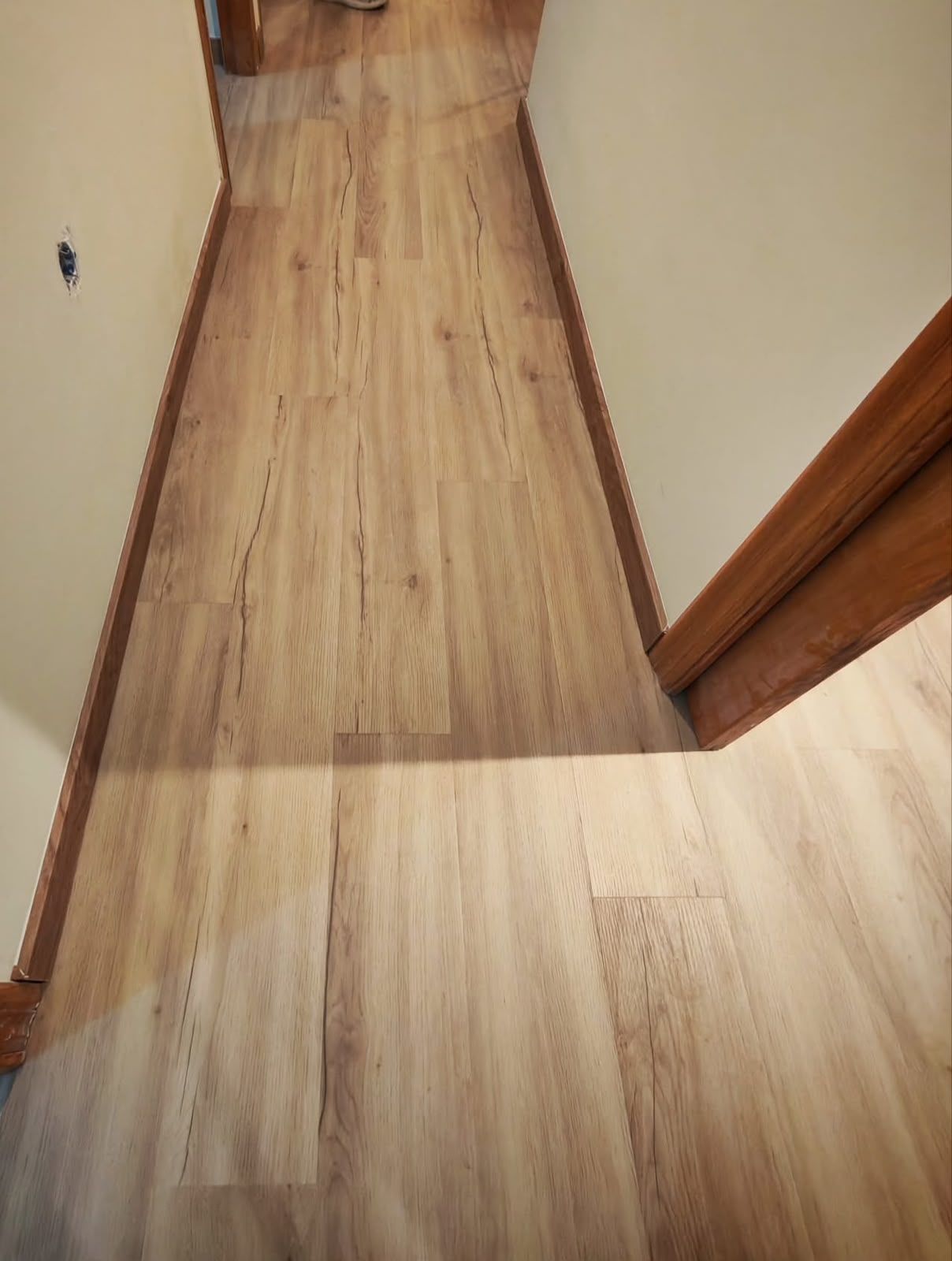 Pasillo estrecho con suelo de madera y paredes beige, que muestra la transición entre distintos acabados del suelo.