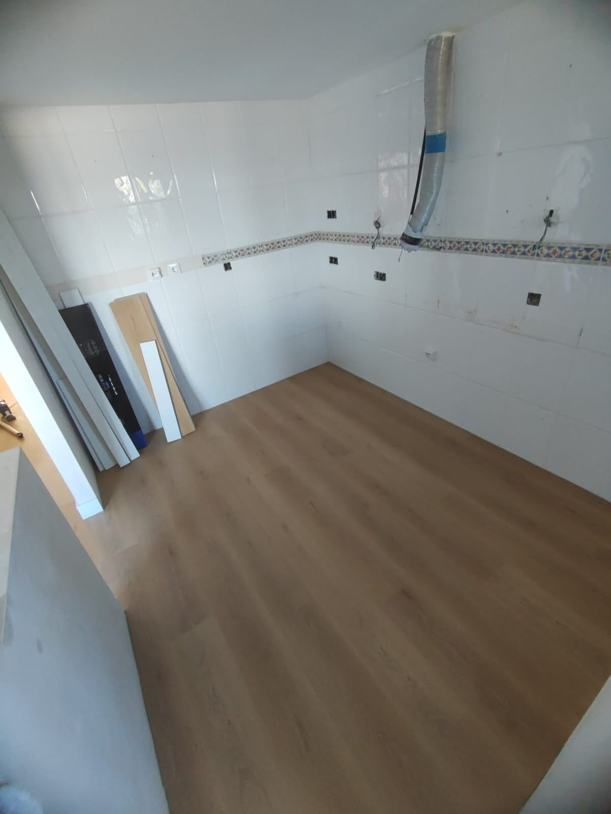 Cocina blanca vacía en proceso de renovación, con suelo de madera y tuberías a la vista en las paredes