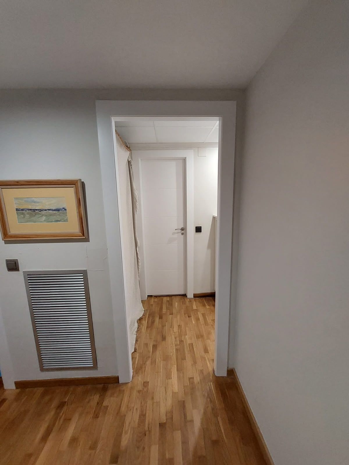 Pasillo estrecho con suelo de madera, paredes grises y una puerta blanca que da a una habitación más luminosa.