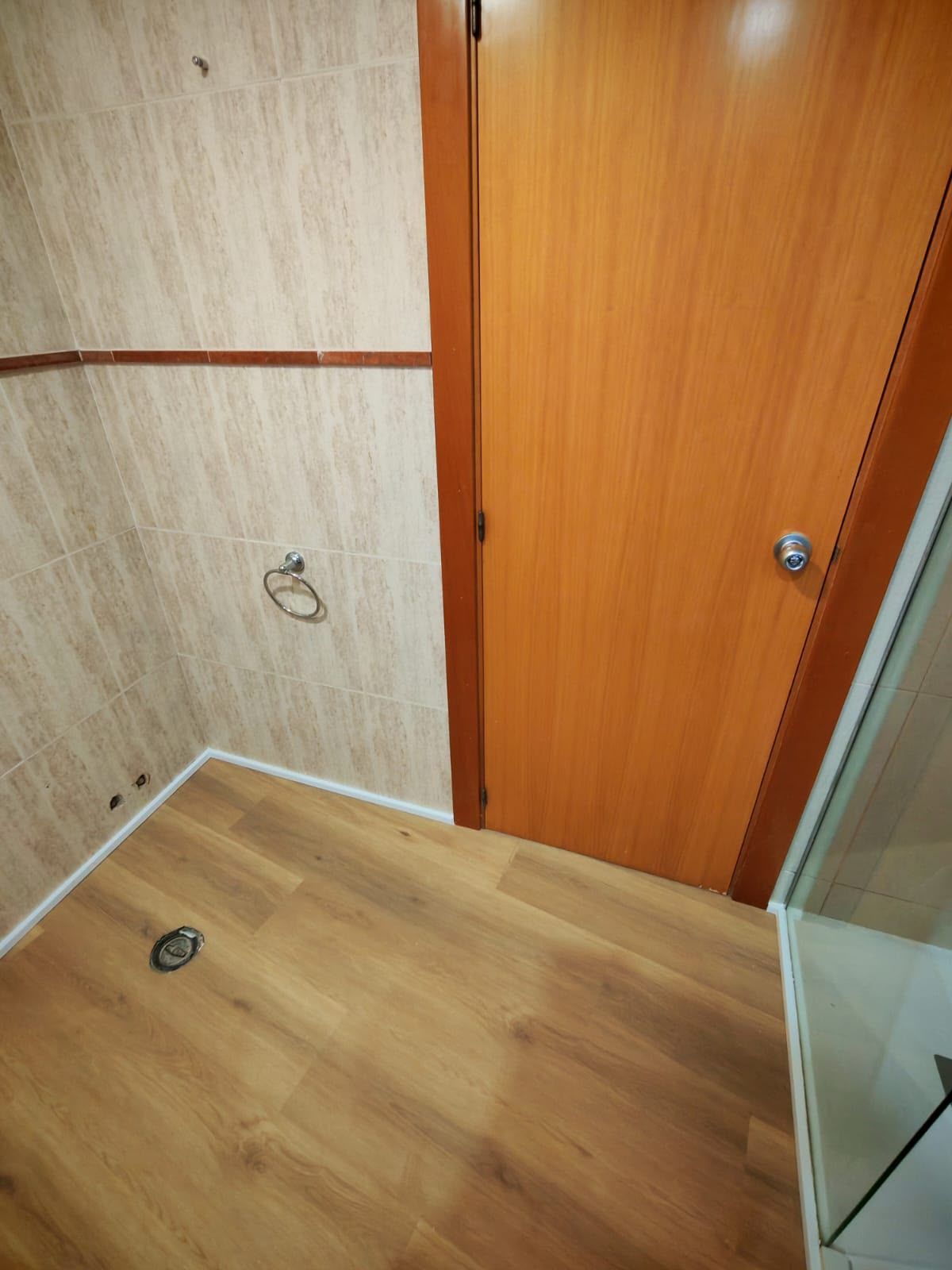 Esquina de un baño pequeño con suelo de imitación madera, pared beige, puerta naranja y mampara de ducha de vidrio.