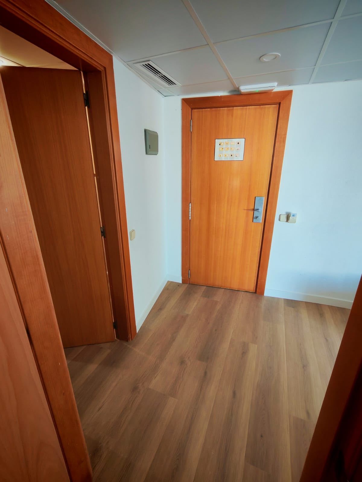 Pasillo estrecho con suelo de madera clara, paredes blancas y puertas naranjas al fondo y en el lado izquierdo