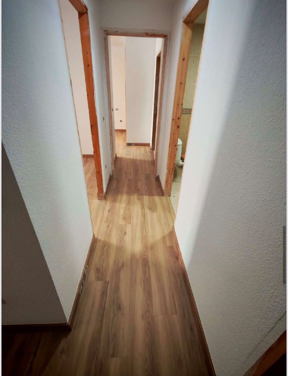 Pasillo estrecho con paredes blancas, suelo de madera y varias puertas abiertas que dan a las habitaciones del fondo