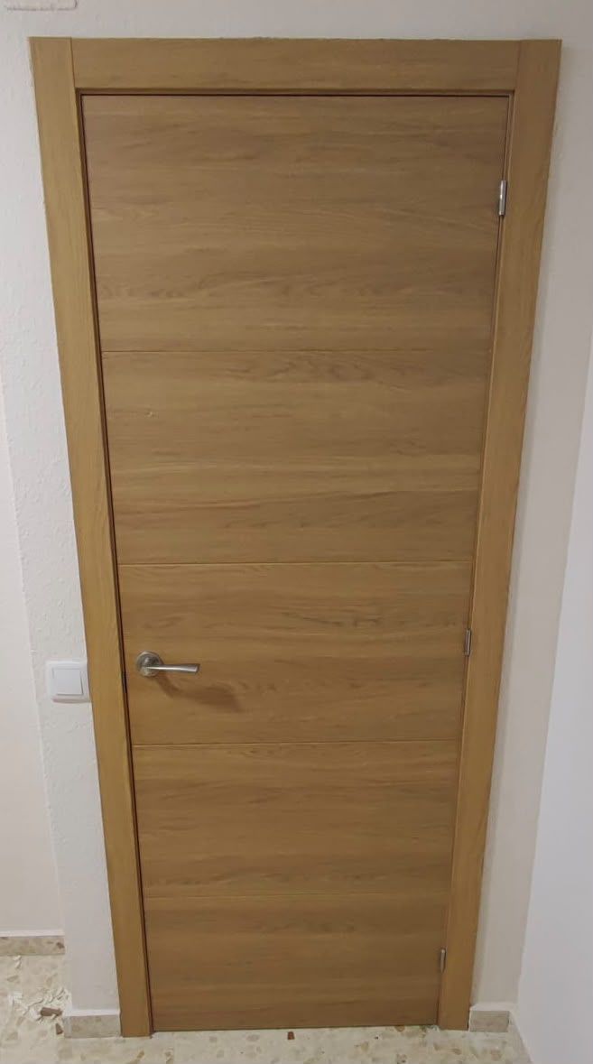 Puerta interior de madera clara cerrada, con manilla plateada, en un hueco de una pared lisa