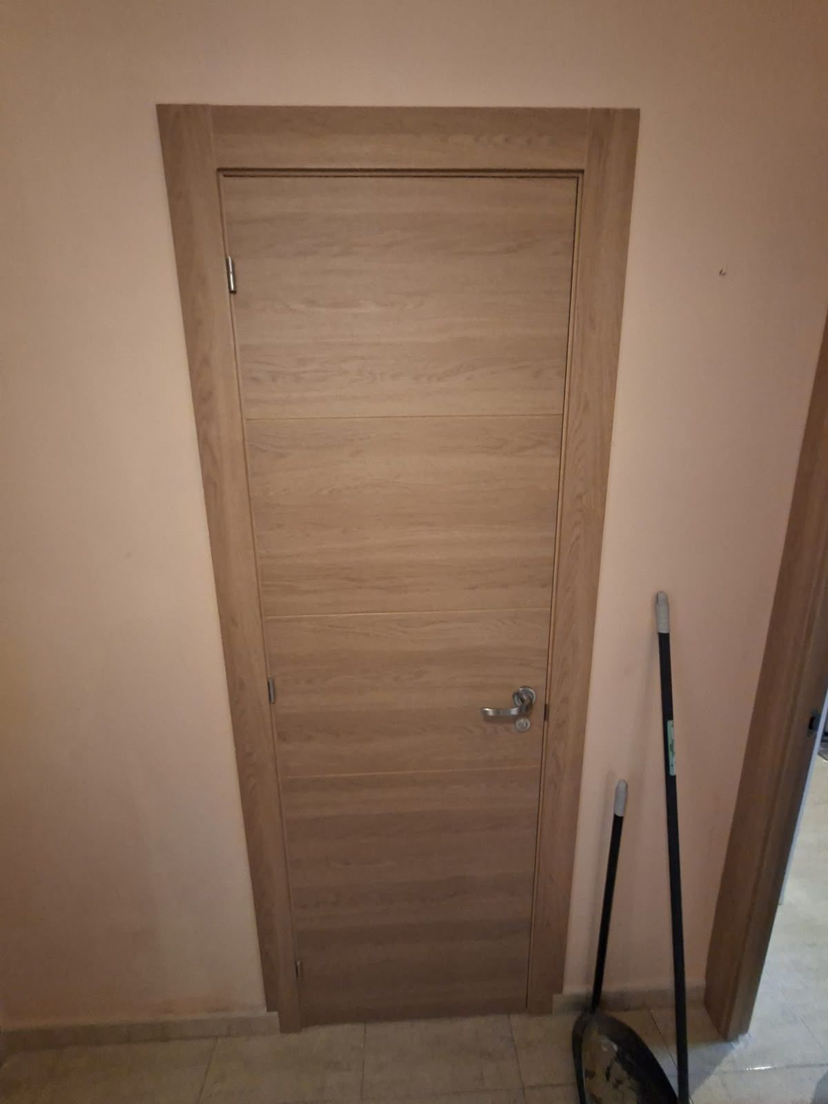 Puerta interior cerrada de madera clara con manilla plateada, junto a una pared y utensilios de limpieza.
