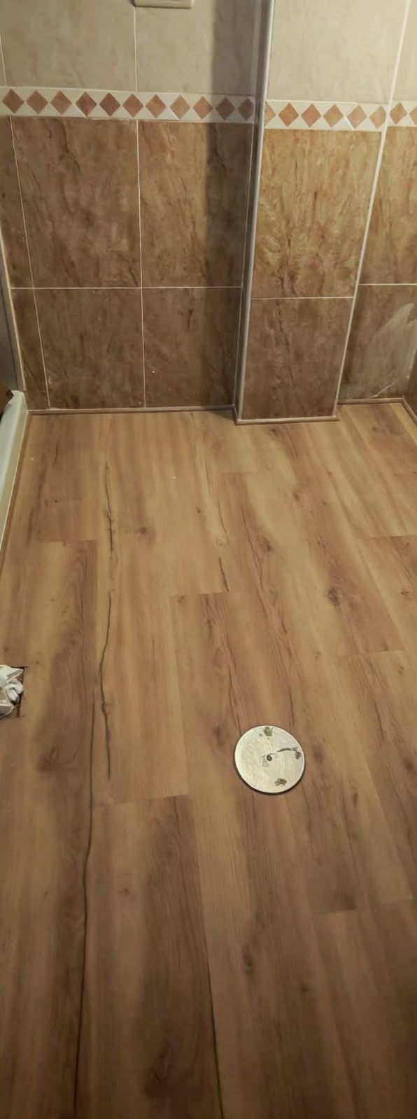 Suelo con aspecto de madera y un desagüe redondo blanco cerca de una pared alicatada.