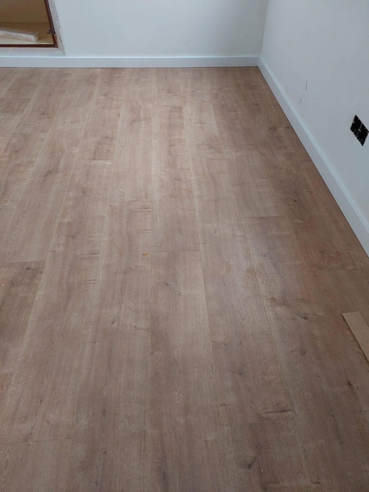 Habitación vacía con suelo de madera clara y paredes blancas, donde se ve una esquina y el zócalo.