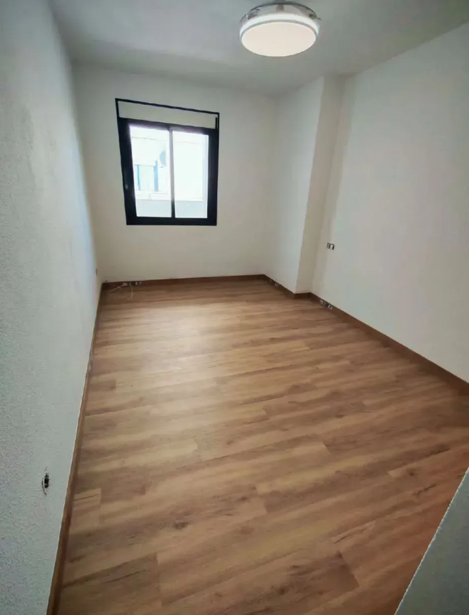 Habitación vacía con suelo de madera, paredes blancas, una lámpara de techo y una ventana con marco oscuro.