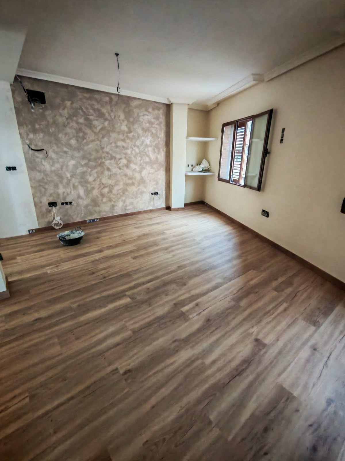 Habitación vacía con suelo de aspecto amaderado, pared decorativa con motivos y una ventana a la derecha.