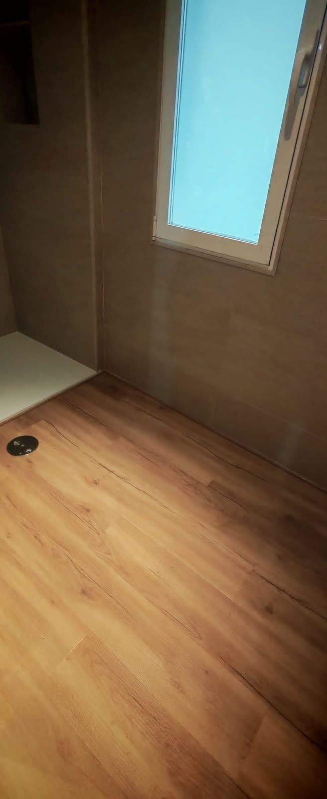 Habitación vacía con suelo de madera y paredes marrones, con una pequeña ventana cerca del techo.