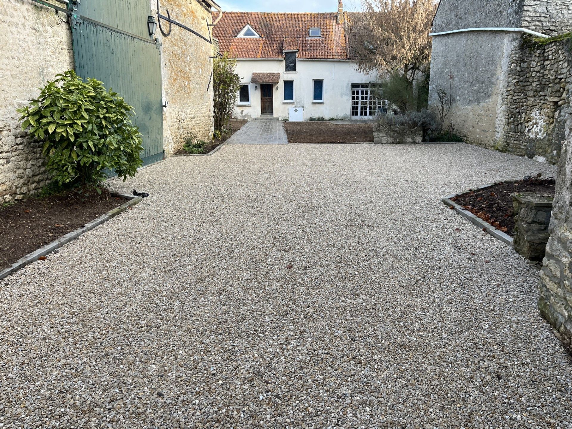 Cour de gravier menant à une maison blanche encadrée de murs de pierre et de verdure.
