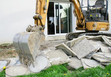 Godet d'excavatrice brisant des dalles de béton devant une maison dont la baie vitrée est ouverte.