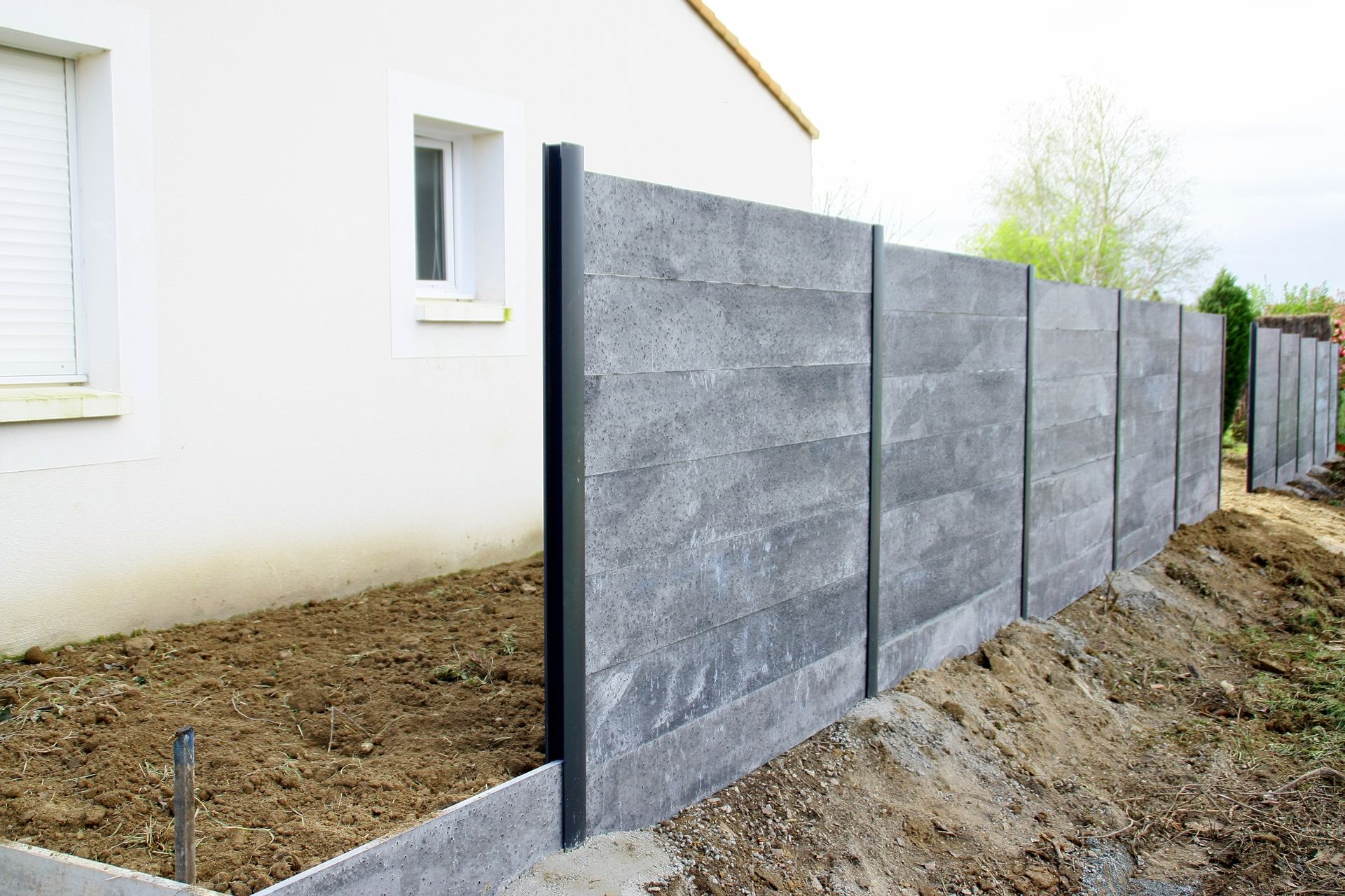 Clôture en panneaux béton gris, installée le long d'une maison dont le jardin est en cours d'aménagement.