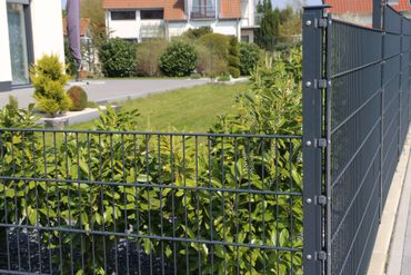 Clôture grillagée en métal noir borde un jardin verdoyant avec des haies et des maisons en arrière-plan.