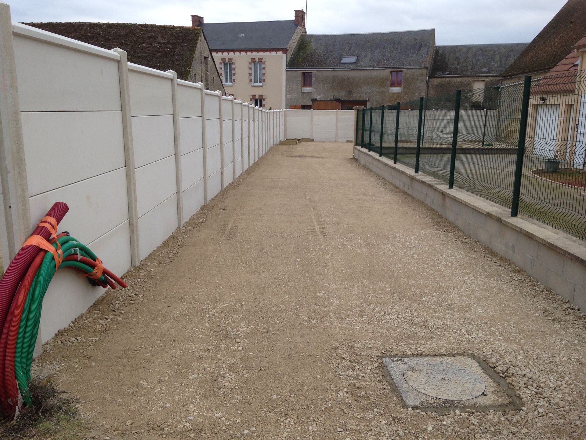 Allée de gravier longeant une clôture blanche, avec des gaines à gauche et une clôture à droite.