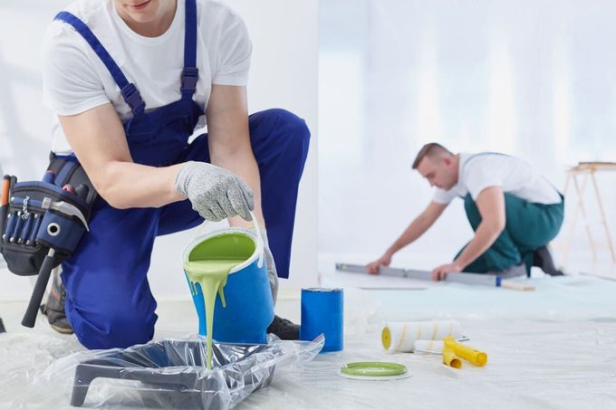 Un pintor vierte pintura verde en una bandeja; otra persona usa un nivel en el piso, ambos en una habitación durante las renovaciones.