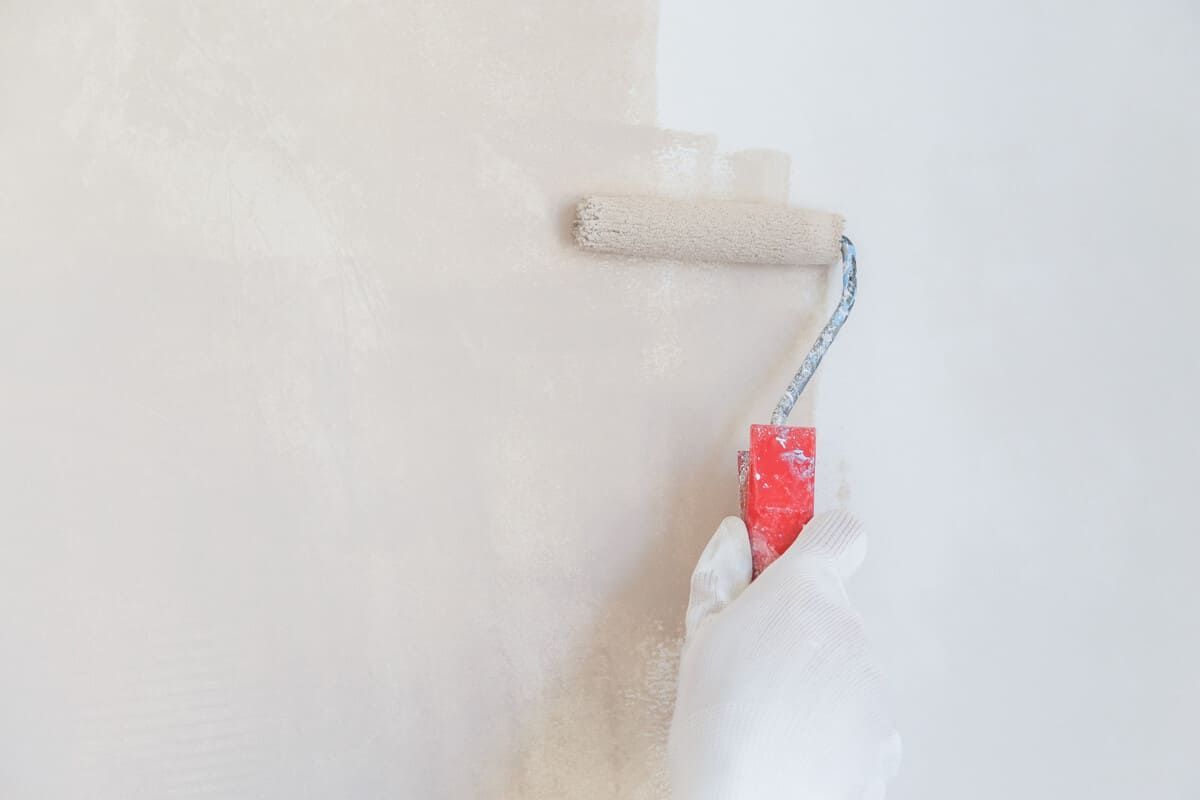Mano con guante blanco pintando una pared con un rodillo de pintura; pintura blanca visible en la pared.