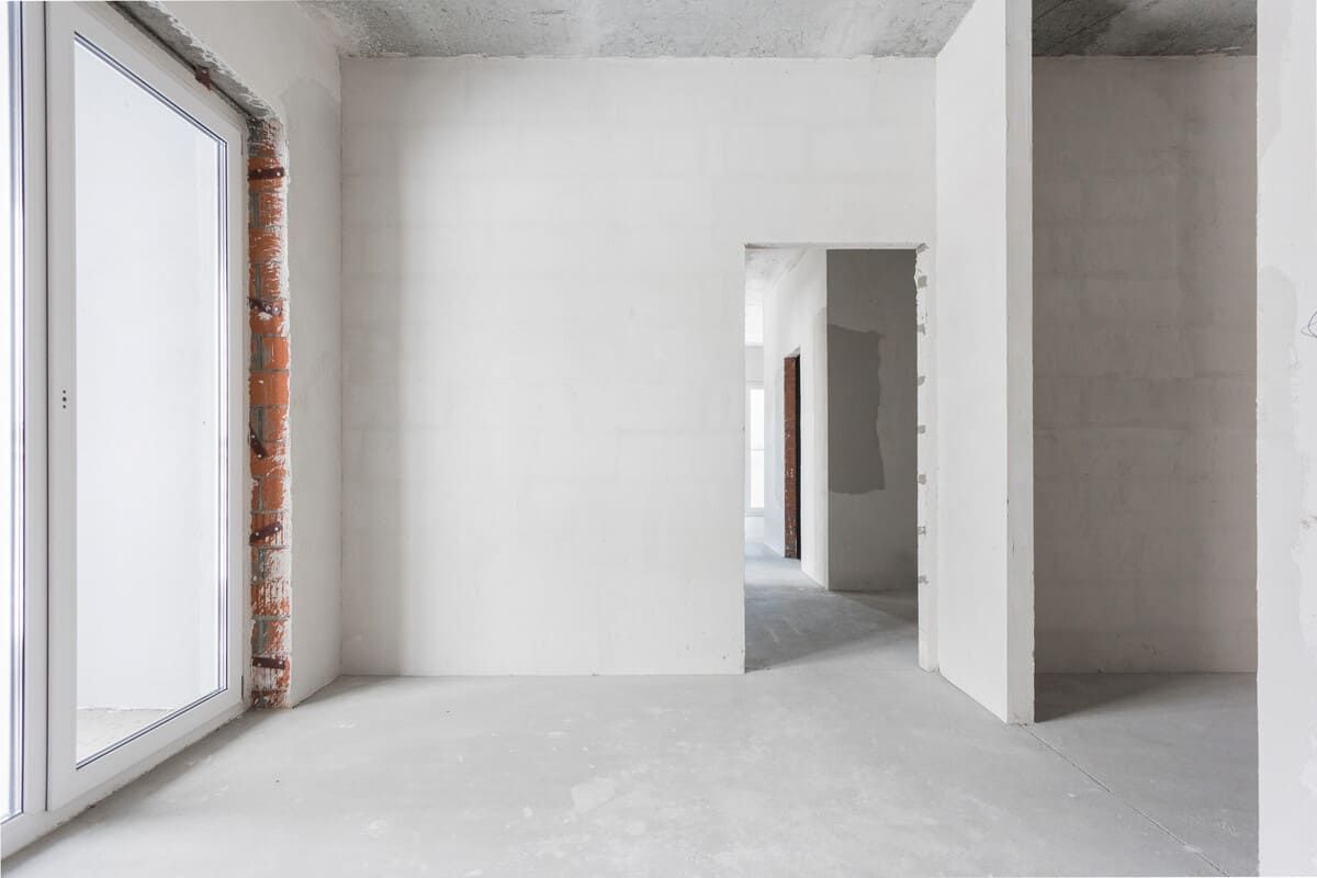 Habitación vacía en construcción; paredes blancas, piso de concreto, ladrillo expuesto alrededor del marco de la ventana.